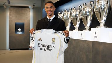 Trent Alexander-Arnold stands holding his new Real Madrid shirt in front of Champions League trophies at Real's training ground